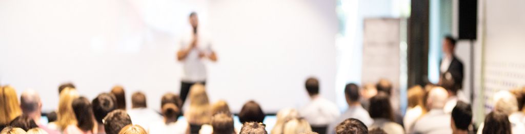 A blurred view of a speaker giving a talk to a room full of people sitting down