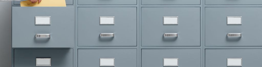 A large grey filing cabinet. A hand reaches into one of the drawers to pull a file out.