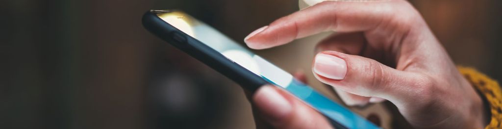 A pair of hands holding a mobile phone. The index finger of the right hand hovers over the lit up screen.