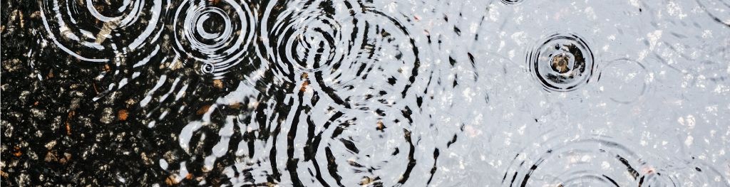 A puddle on a gravelled surface with raindrops hitting it, creating a pattern of concentric circular ripples on its surface.