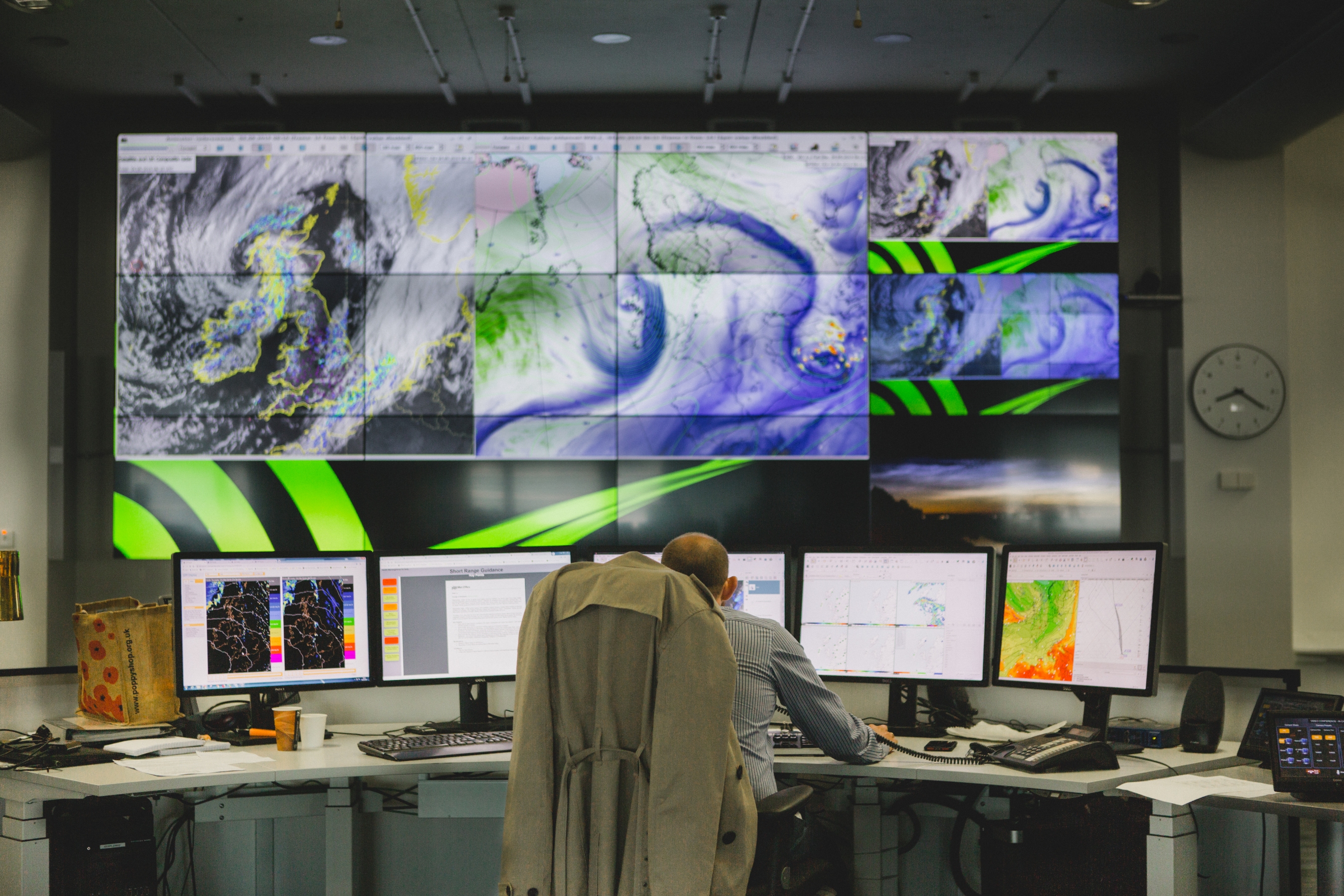 This photo features a large computer screen showing a weather system. A person is sat at a desk in front of it.