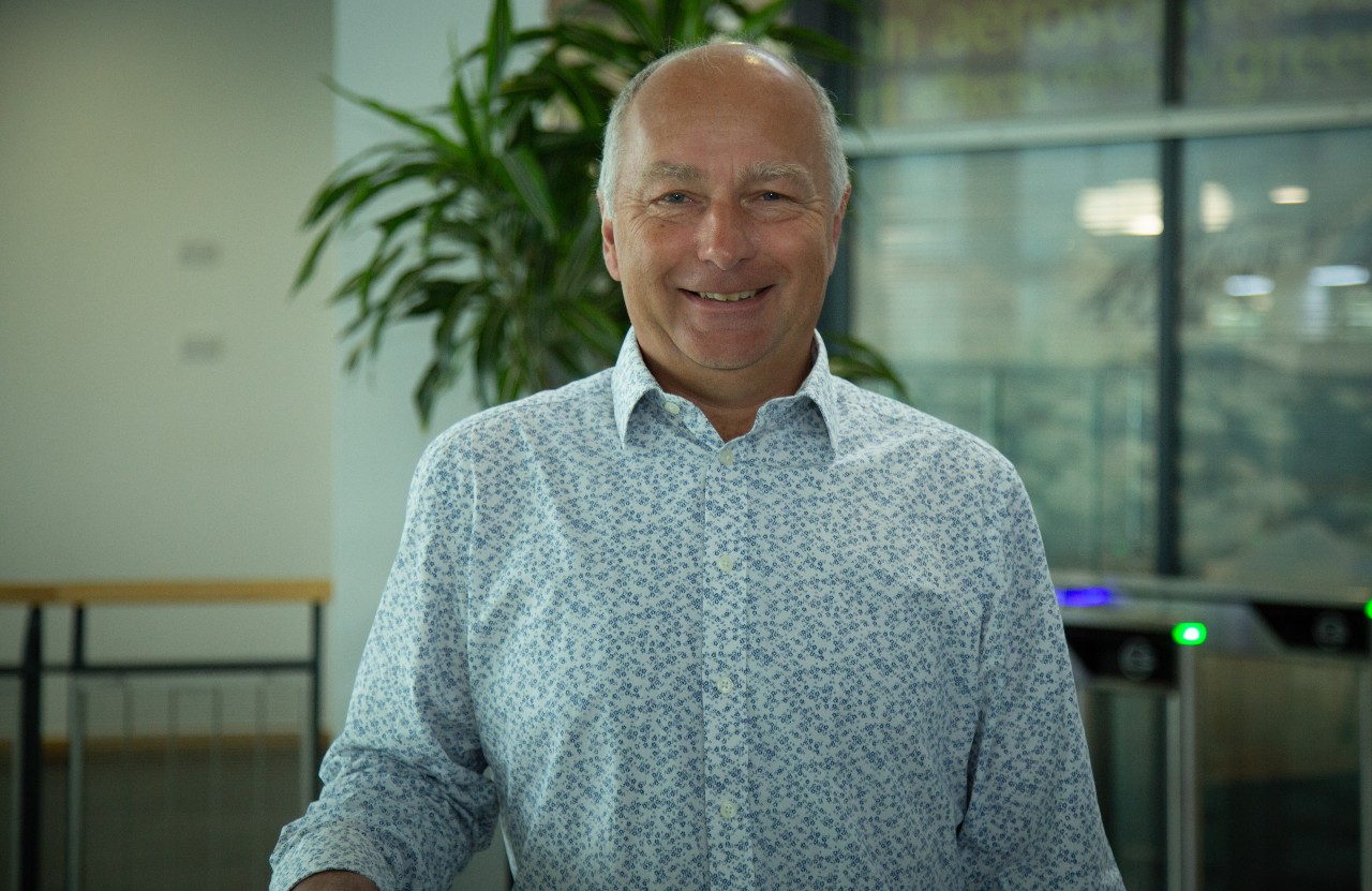 Professor Peter Stott MBE, Climate Science Fellow at the Met Office standing in the Met Office HQ for a portrait photo wearing a patterned shirt with a plant in the background.