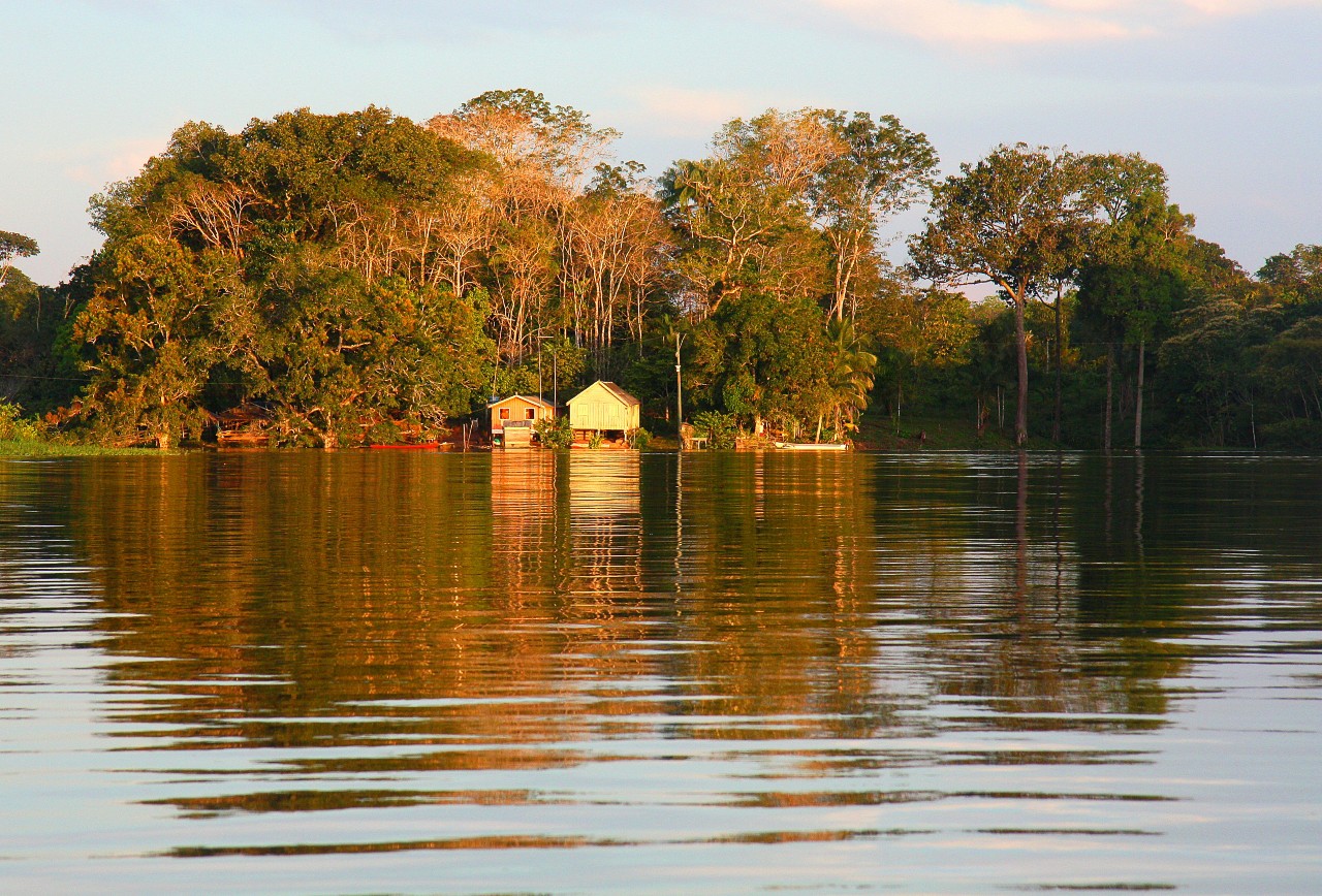 Calm river in the foreground with huts on the shoreline in the background and a forest behind them with blue skies above.